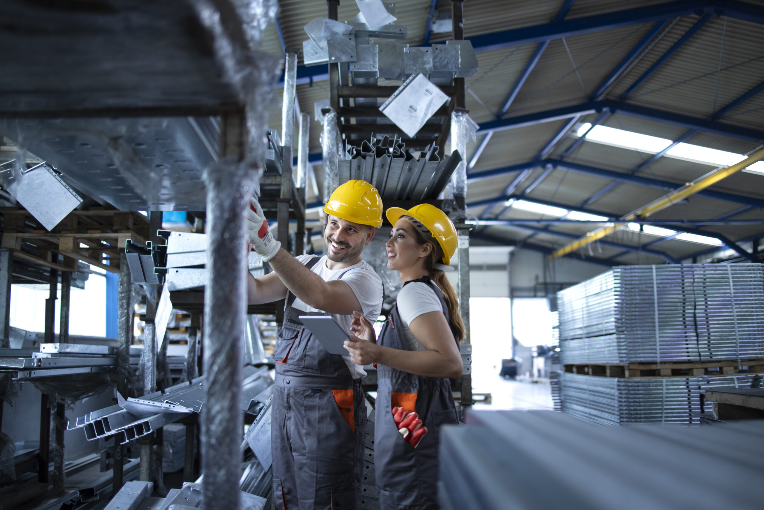 Factory workers checking inventory with tablet computer in industrial warehouse full of metal parts.